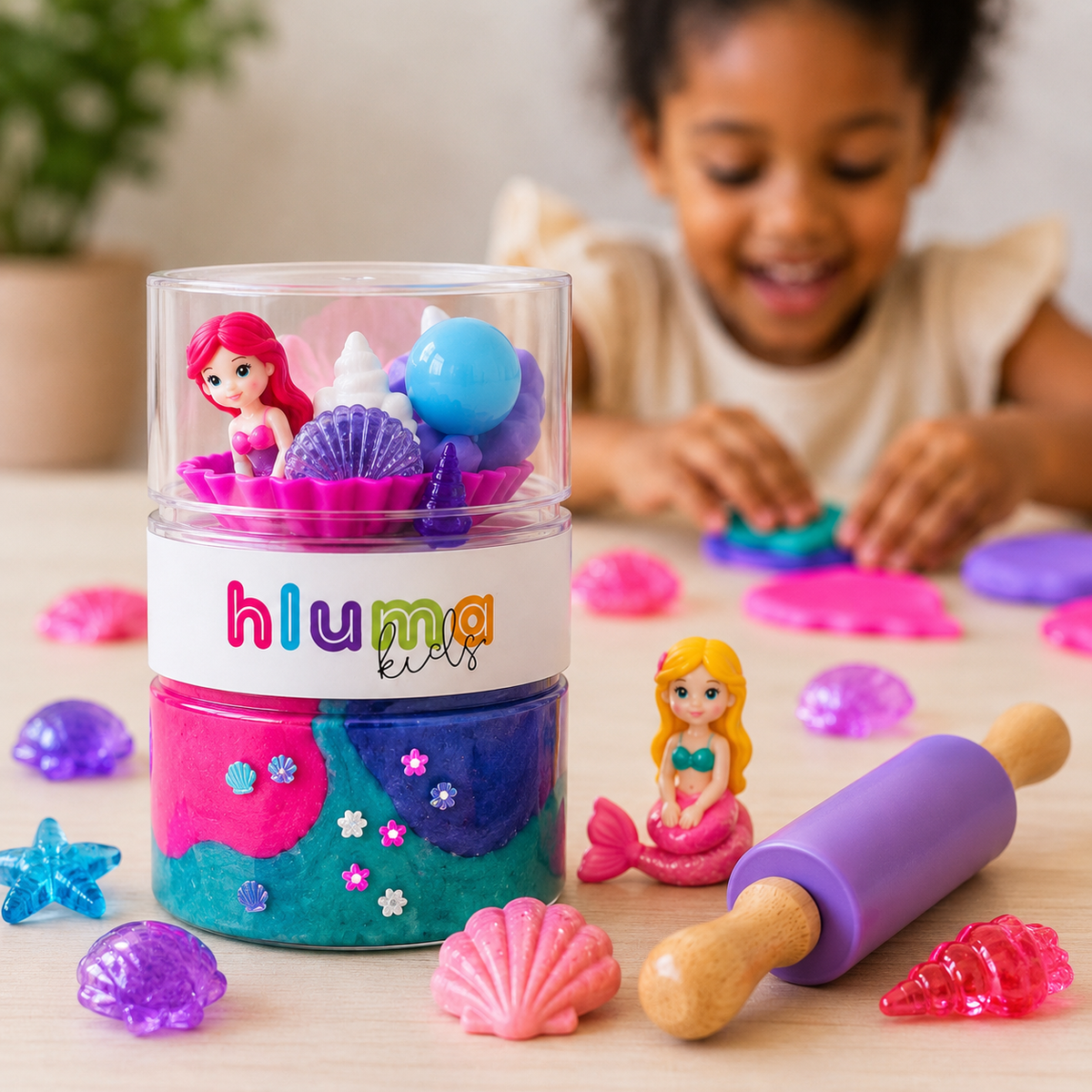 Close-up of a Hluma Kids Mermaid Lagoon Wonder Jar with a mermaid figure and shell pieces, a child playing with pink playdough in the background